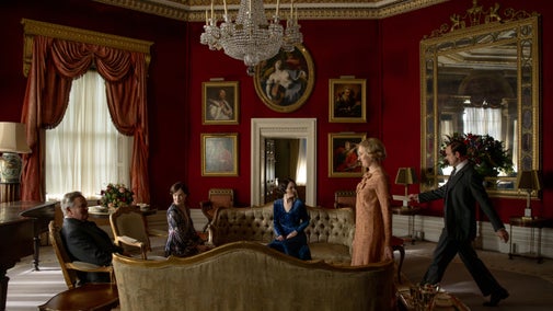 Some of the cast of Downton Abbey sitting on sofas in a circle with two members walking to join them in an ornate room, with red walls and many paintings around them.